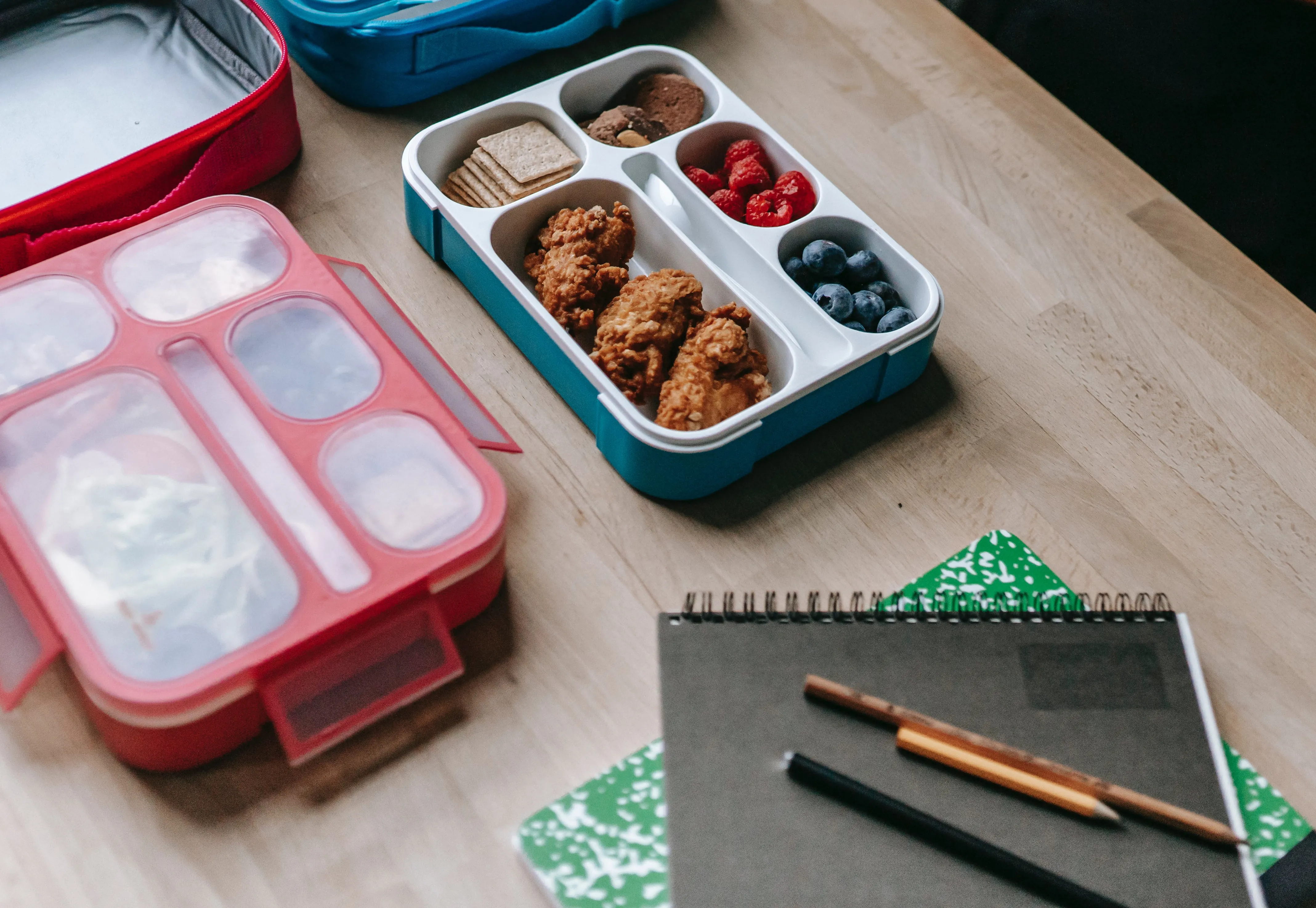Several lunch containers with food in them on a table with piled notebooks